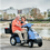 Couple Driving Dual Seat Recreational Scooter On Boardwalk.